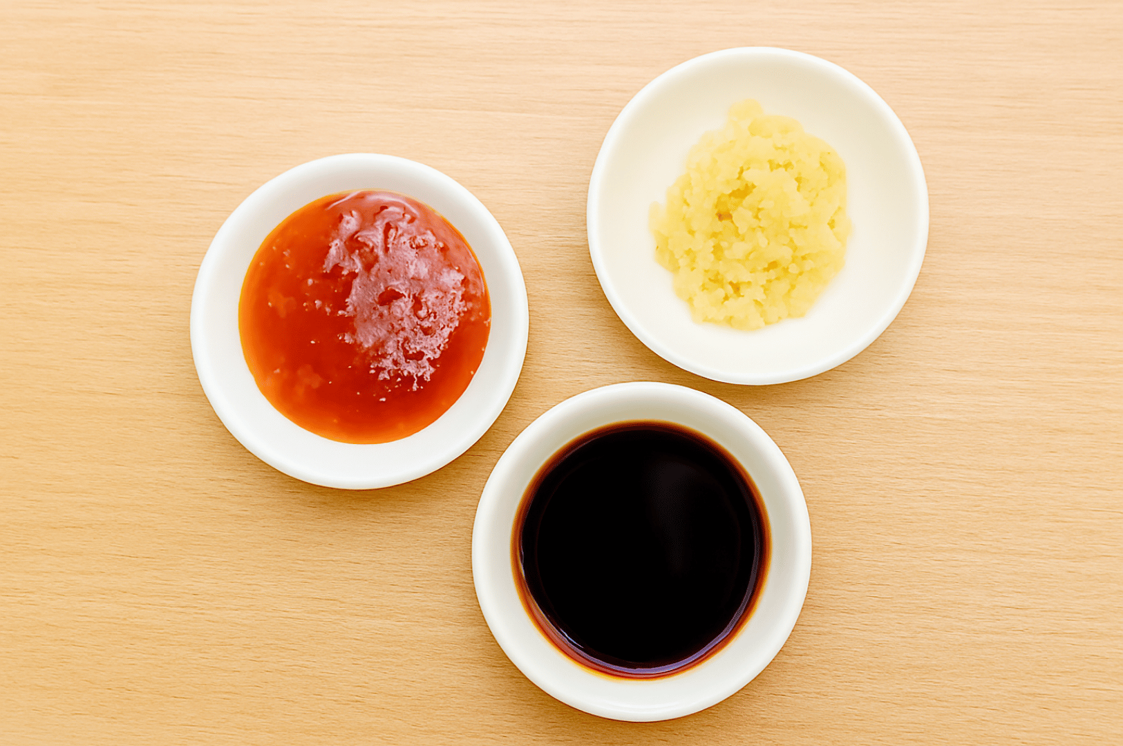 Hainanese-Chicken- (4) Three white bowls on a light wooden surface, each holding a key flavor: glossy sweet chili sauce, finely minced garlic, and dark soy sauce—an understated trio that anchors the sensory depth of Hainanese Chicken Rice.