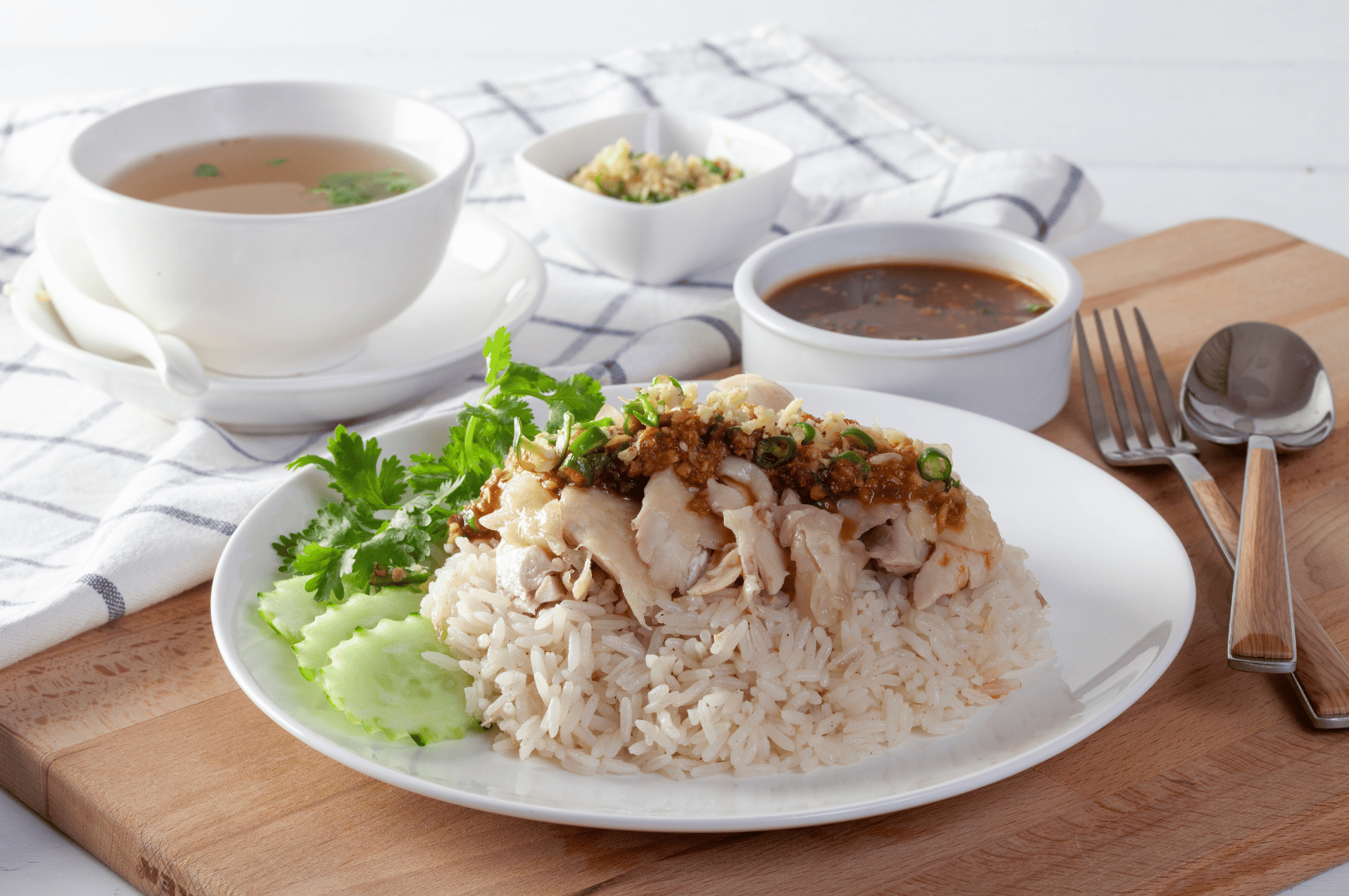 Hainanese-Chicken- (5) Poached chicken slices resting on white rice, garnished with fresh cilantro and cucumber, flanked by clear herb soup, dark dipping sauce, and minced ginger—an intimate, home-style presentation that distills the comfort and quiet sophistication of Hainanese Chicken Rice.