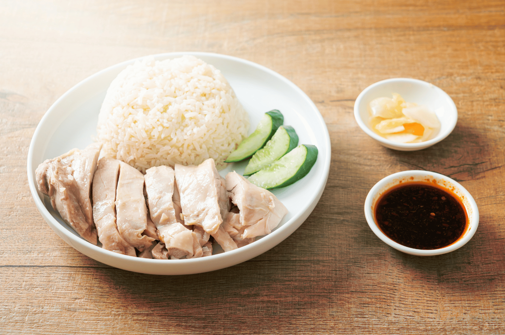 Hainanese-Chicken- (8) Neatly arranged poached chicken over seasoned rice, garnished with cucumber slices and paired with dark soy sauce and pickled ginger—an understated composition that distills the harmony and refined simplicity of Hainanese Chicken Rice
