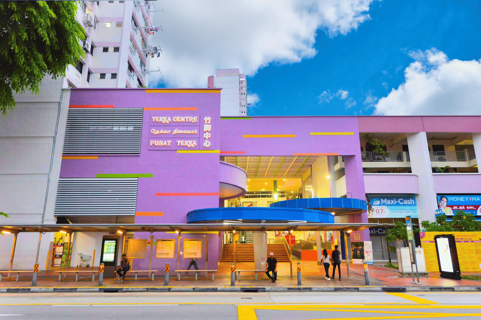 Little-India-Food- (2) Exterior of Tekka Centre in Little India, Singapore—its bold purple façade and multilingual signage signal a vibrant crossroads of cultures, flavors, and stories. A gateway to the district’s spice-scented food stalls with rice cakes and chicken rice and other ingredients from north india and middle east traditionally eaten with the bustling market life, captured under a bright tropical sky.