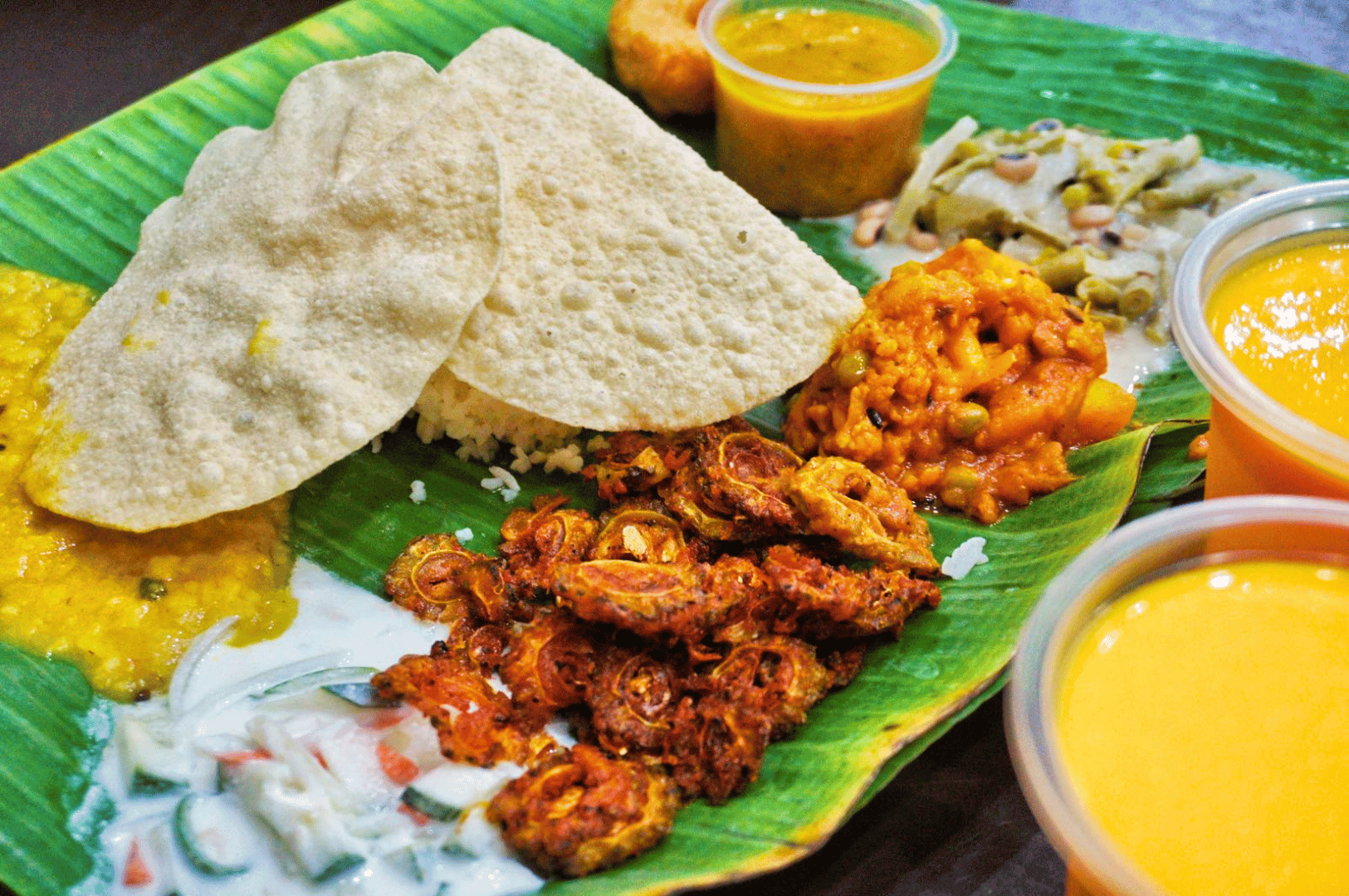 Little-India-Food- (3) Traditional South Indian banana leaf meal served in Little India, Singapore—an aromatic mosaic of crispy papadum, creamy dal, tangy raita, spiced vegetable fritters, and vibrant curries, arranged with ceremonial care. A sensory feast that captures the heart of the Spice Route in every bite.