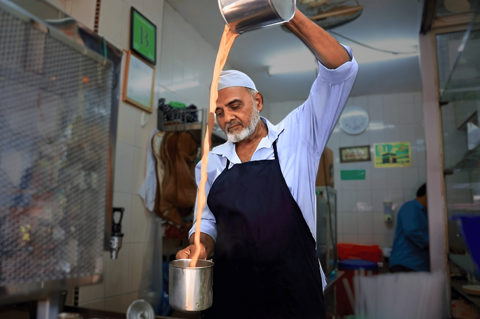 Little-India-Food- (5) Teh tarik master in action—streaming hot pulled tea between metal vessels with theatrical flair in a Little India kitchen. His precise movements and focused expression capture the artistry behind this beloved beverage ritual, steeped in spice, warmth, and community.