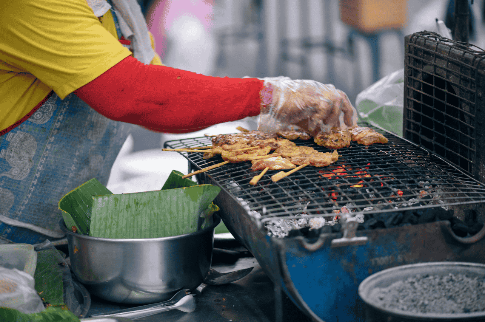The-Best-Satay-in-Singapore-hawker Street food vendor grilling skewers of well-marinated satay over glowing charcoal, capturing the smoky aroma and flame-kissed char that define the best satay in Singapore. Wearing a yellow shirt and blue apron, the vendor prepares juicy chicken satay, pork satay, beef satay, mutton satay, and pork belly satay with traditional techniques at iconic satay stalls like Chai Ho Satay, Chuan Kee Satay, Shi Xiang Satay, Ah Pui Satay, Pang's Satay, and Yong Seng Satay. Found in hawker centres such as Lau Pa Sat, Newton Food Centre, and Chinatown Complex Market Food Centre at 335 Smith Street, these satay sticks are grilled to perfection and served with rich satay sauce made from peanuts, pineapple puree, grated pineapple, and subtle hints of lemongrass, coriander, and cumin. The scene evokes the vibrant energy of satay street culture, where generous portions of lean meat, beef tripe, and lamb are cooked with care, delivering a spicy, savoury, and sweet bite that celebrates old school satay and modern Malay style satay at an affordable price