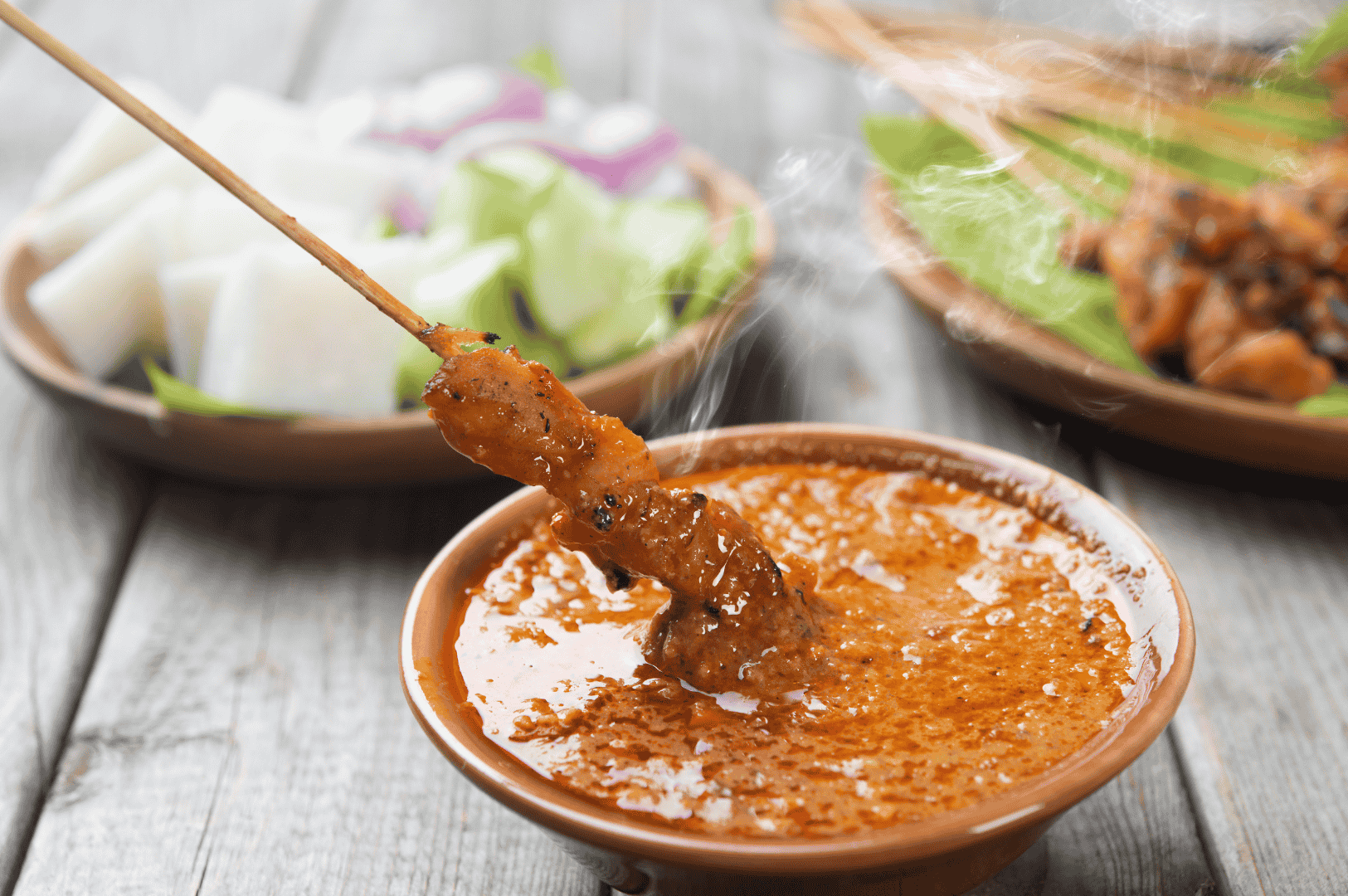 The-Best-Satay-in-Singapore-magic Close-up of juicy grilled chicken satay dipped into thick, spicy peanut sauce, showcasing the best satay in Singapore. The satay stick glistens with a charred exterior and well-marinated meat, surrounded by traditional sides like lontong rice cakes, cucumber, and red onion. This image evokes the smoky aroma and savoury sweetness found at iconic satay stalls across hawker centres like Lau Pa Sat, Newton Food Centre, and Chinatown Complex Market Food Centre. Whether it’s pork satay, pork belly satay, beef satay, mutton satay, or even beef tripe, Singapore’s satay scene—from Chai Ho Satay and Chuan Kee Satay to Shi Xiang Satay and Ah Pui Satay—offers generous portions of tender, lean meat grilled to perfection. The satay sauce, rich with peanuts, pineapple puree, and subtle hints of lemongrass, coriander, and cumin, delivers a flavourful bite that defines old school satay and modern Malay style satay alike.