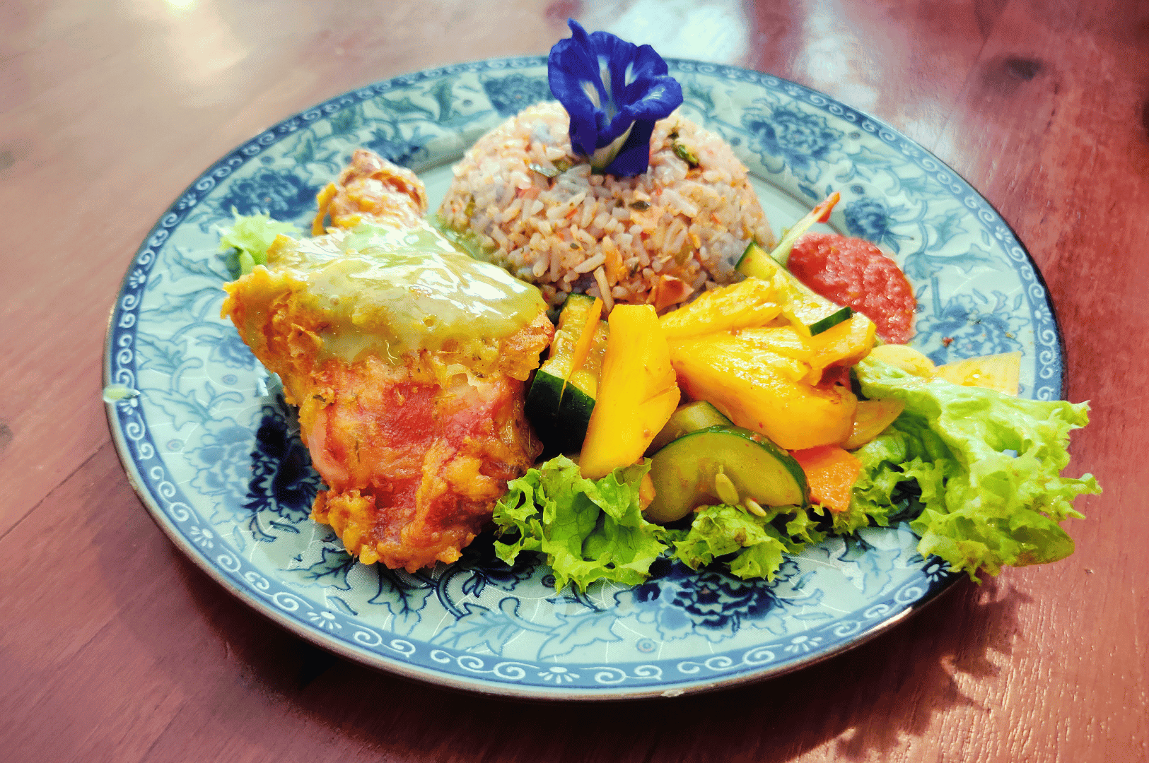 Chinese-Malay-Fusion- (6) Artfully plated Chinese-Malay fusion dish on a blue-and-white ceramic plate: golden herb-crusted chicken, tropical mango and cucumber medley, and fragrant fried rice crowned with a vivid blue edible flower—an elegant composition that channels Peranakan refinement and Nyonya vibrance, challenging Singapore’s culinary boundaries with grace and defiance.