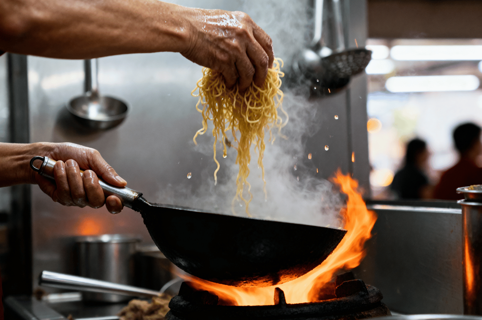 Hawker-Photo- (2) Hands poised over a blazing wok as a hawker chef lifts a tangle of yellow noodles into the heat, framed by rising steam and the glow of open flame. The scene captures the tactile choreography and sensory intensity of Singapore’s hawker kitchens, where every gesture speaks to tradition, speed, and mastery.