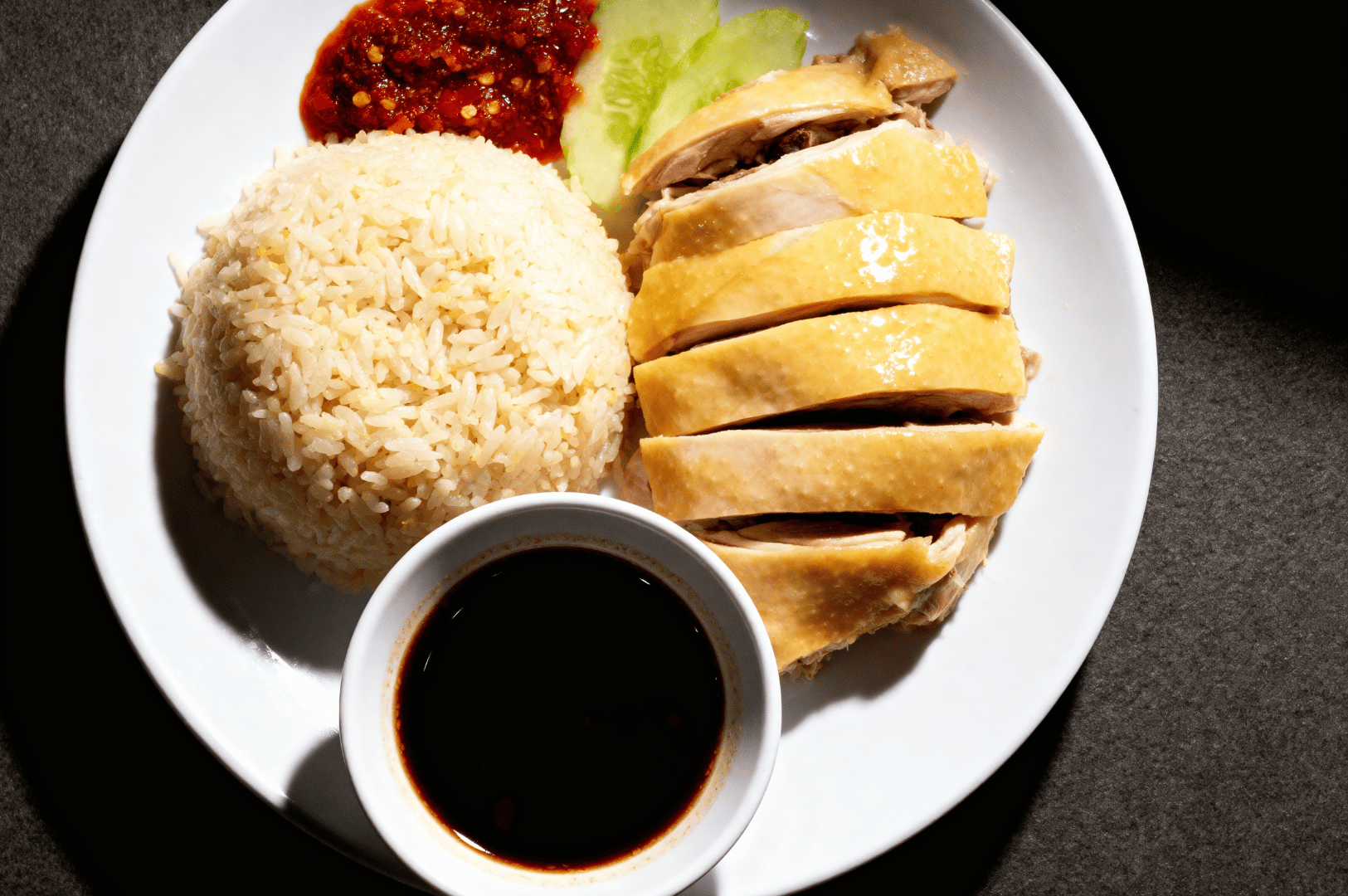 Steam-and-Shadows- (3) A pristine plate of Hainanese chicken rice sits under soft lighting, its elements arranged with quiet precision—poached chicken glistening with golden skin, fragrant rice molded into a neat mound, and vibrant chili sauce dotted with seeds beside crisp cucumber slices. A small bowl of dark soy sauce anchors the composition. The dish radiates understated elegance, offering a moment of stillness amid the fire and motion of the hawker kitchen. It’s the final beat in the dance—where heat gives way to harmony, and Singapore’s culinary soul is served, one plate at a time.