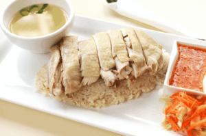 Poached Hainanese chicken sliced and arranged over fragrant rice, paired with clear chicken broth, fiery red chili sauce, and pickled vegetables—an elegant composition capturing the quiet allure and cultural depth of Singapore’s iconic comfort dish.