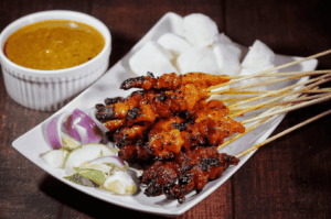 A plate of grilled meat skewers featuring chicken satay, pork satay, mutton satay, and beef satay served with satay sauce and thick peanut sauce, alongside rice cakes and sliced cucumbers and onions—showcasing the best satay in Singapore. Captured at a hawker centre like Chinatown Complex Market Food Centre or Newton Food Centre, this satay in Singapore highlights tender meat, charred exterior, juicy bite, and well-marinated flavours with subtle hints of lemongrass, coriander, cumin, and pineapple puree. Popular satay stalls such as Chai Ho Satay, Chuan Kee Satay, Yong Seng Satay, Shi Xiang Satay, Ah Pui Satay, Pang's Satay, and Chomp Satay at 335 Smith Street and Satay Street offer generous portions of pork belly satay, beef tripe, lamb, prawns, and lean meat skewers. Served on five sticks at an affordable price, this old school satay delivers sweet, savoury, spicy taste and oily richness with grated pineapple and peanuts—an iconic malay style satay experience at Lau Pa Sat and other food centres.