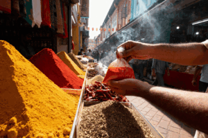 A bustling spice market in Little India, where vibrant mounds of turmeric, chili, and cumin spill from metal trays along a narrow street. In the foreground, a hand holds a small bag of fiery red chili powder, while aromatic smoke drifts upward, evoking the heat and fragrance of the spices. The scene captures the sensory seduction of the spice trail—color, scent, and movement converging in a vivid portrait of culinary tradition.