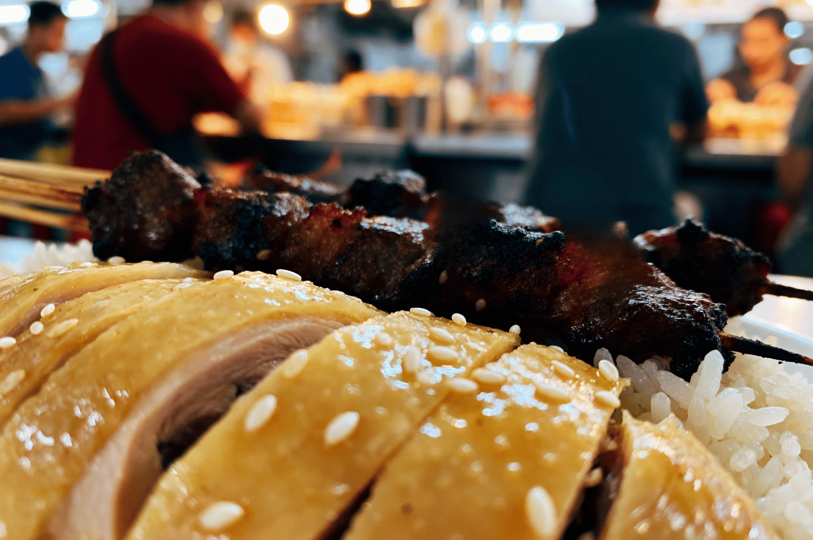 Macro-Photography- (1) Close-up of grilled chicken skewers with rice on a table, captured in dramatic macro style at a Singapore eatery, with blurred diners in the background.