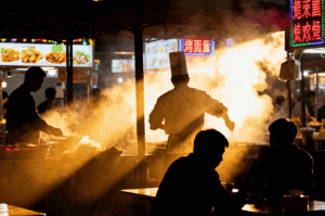 A chef in a tall white hat stands center stage at Maxwell Food Centre, bathed in dramatic backlight as steam swirls around him like smoke in a spotlight. His hands move with precision over sizzling pans, while silhouetted figures behind him prep ingredients in quiet rhythm. Neon signs with Chinese characters glow above the bustling hawker stall, casting vibrant hues across diners seated in the foreground. The interplay of light, shadow, and vapor evokes a theatrical dance—an homage to Singapore’s hawker culture where culinary mastery meets street-side intimacy.