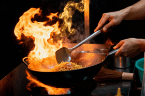 Chef stir-fries Char Kway Teow in a blazing wok, flames erupting dramatically around caramelized noodles and glistening sauce—capturing the elusive wok hei, that smoky, forbidden pleasure at the heart of Singapore’s hawker soul. Shot in warm, high-contrast lighting with a shallow depth of field to emphasize motion and heat.