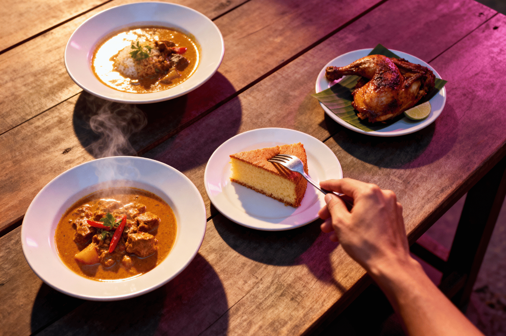 A hand reaches toward a steaming bowl of curry as four vibrant dishes—roasted chicken on banana leaf with lime wedges, spiced meat curries, and a golden slice of cornbread—rest on a wooden table bathed in warm, pink-hued light. The scene captures the convivial heart of colonial dining in Singapore, where Anglo-Indian flavors met Southeast Asian bounty. This moment of indulgence and intimacy reflects the culmination of spice-laden journeys, from maritime trade to shared meals, where history is tasted in every bite.