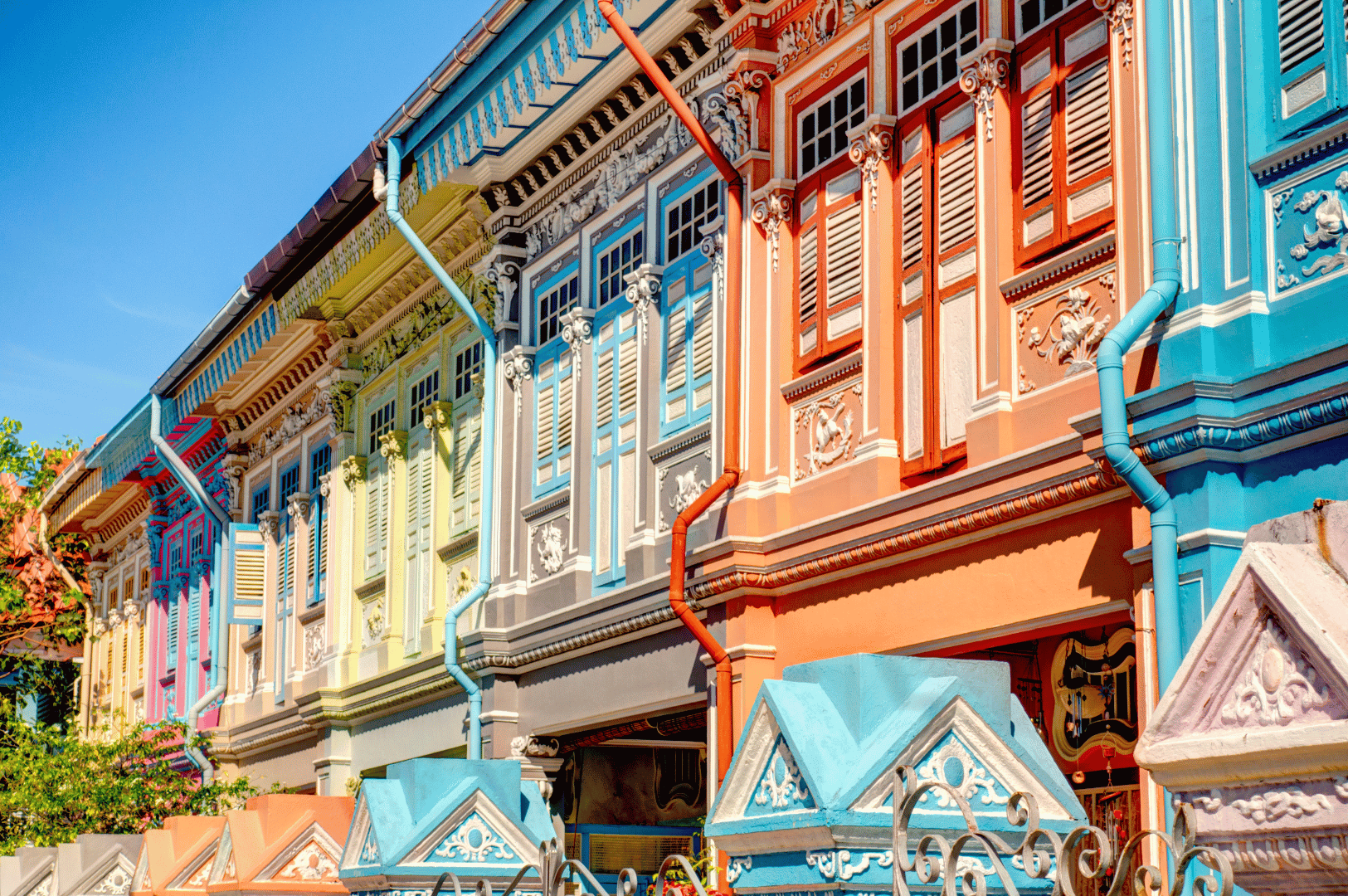 Katong- (2) A vivid row of Katong shophouses in fiery reds, blues, and yellows flaunts intricate Peranakan craftsmanship—carved shutters, ornate moldings, and pastel fencing—evoking the district’s exuberant spirit and its street food pleasures.