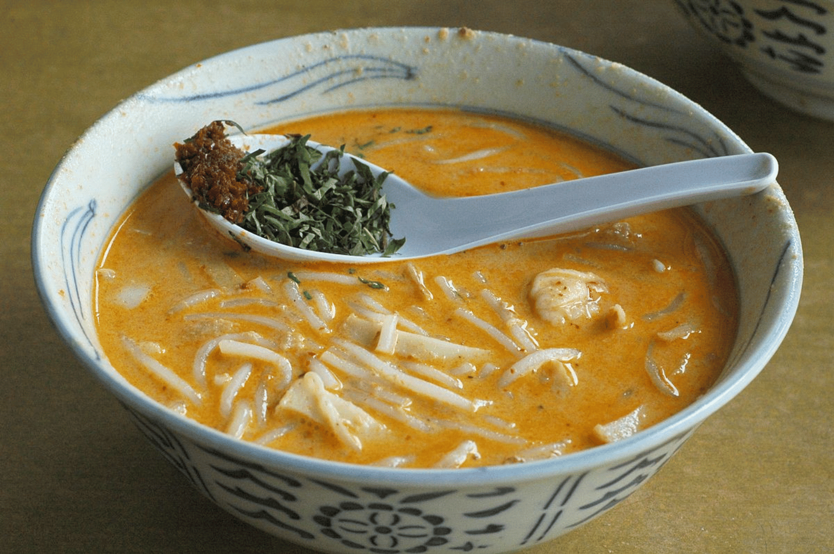 Katong- (3) A bowl of Katong laksa glows with orange coconut curry broth, cradling noodles, tofu, and chili paste—an aromatic embodiment of Peranakan street food indulgence.