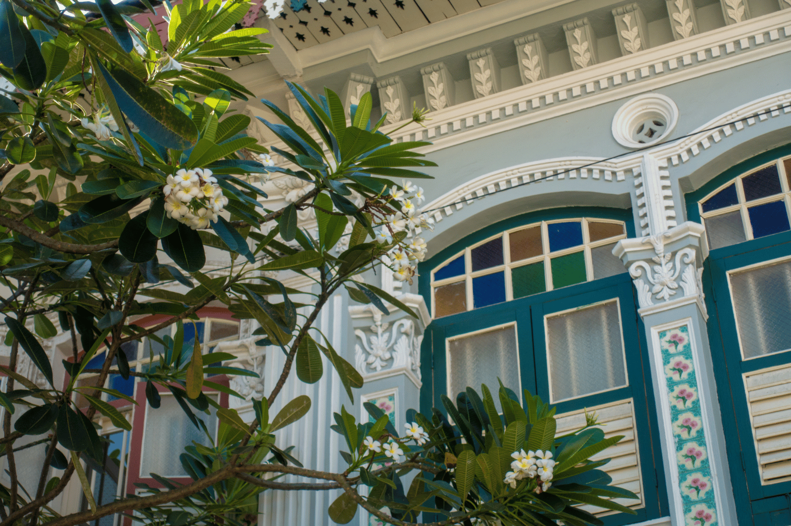 Katong- (5) A close-up of a Katong shophouse facade reveals Peranakan elegance—floral tiles, stained glass, and frangipani blooms—merging architectural artistry with the sensual spirit of street food culture.