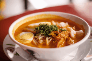 A steaming bowl of Singapore laksa with noodles, prawns, egg, and rich coconut curry on a table.