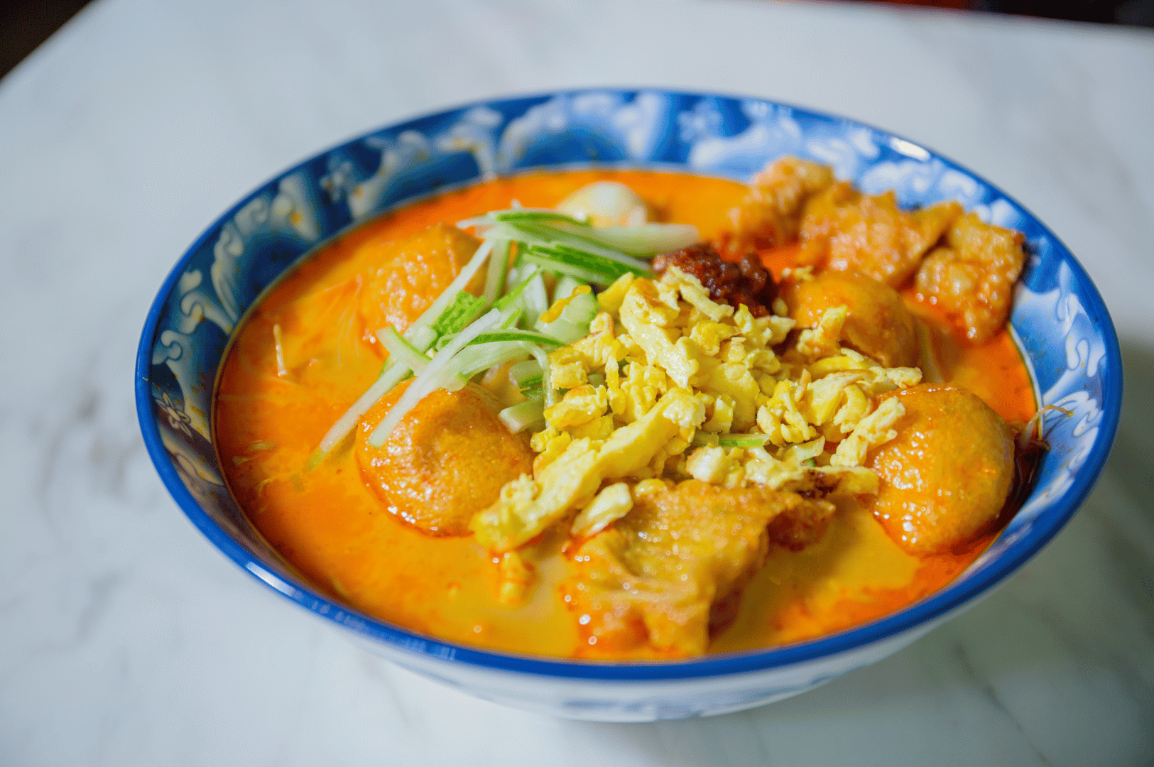 Laksa-Queen- (3) A blue-patterned bowl of creamy laksa with noodles, fish cakes, tofu puffs, and fresh greens on a marble table