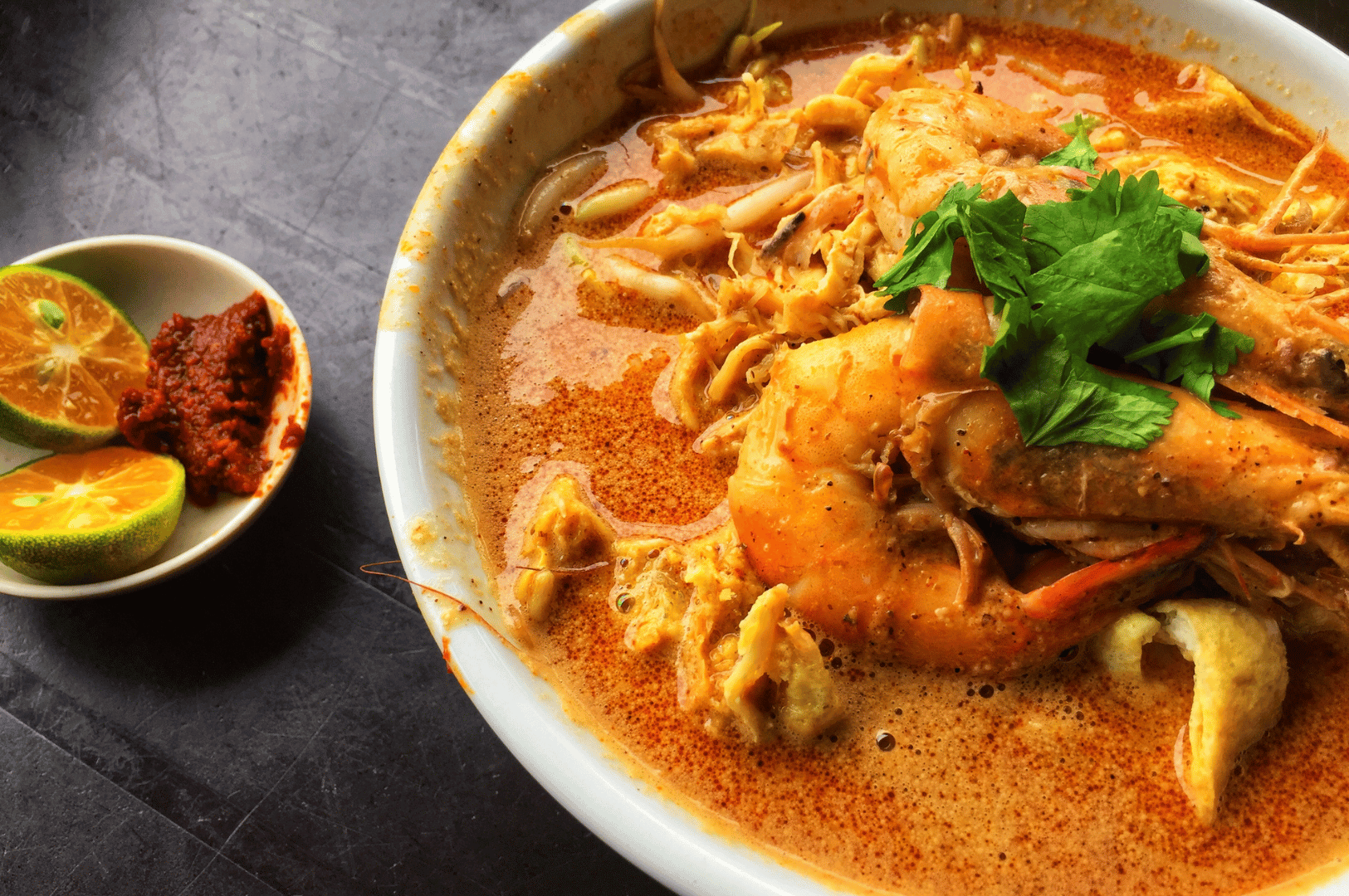 Laksa-Queen- (4) Overhead view of a rich laksa bowl with prawns, noodles, and fresh herbs, with lime halves and sambal on the side—Singapore street food at its best.