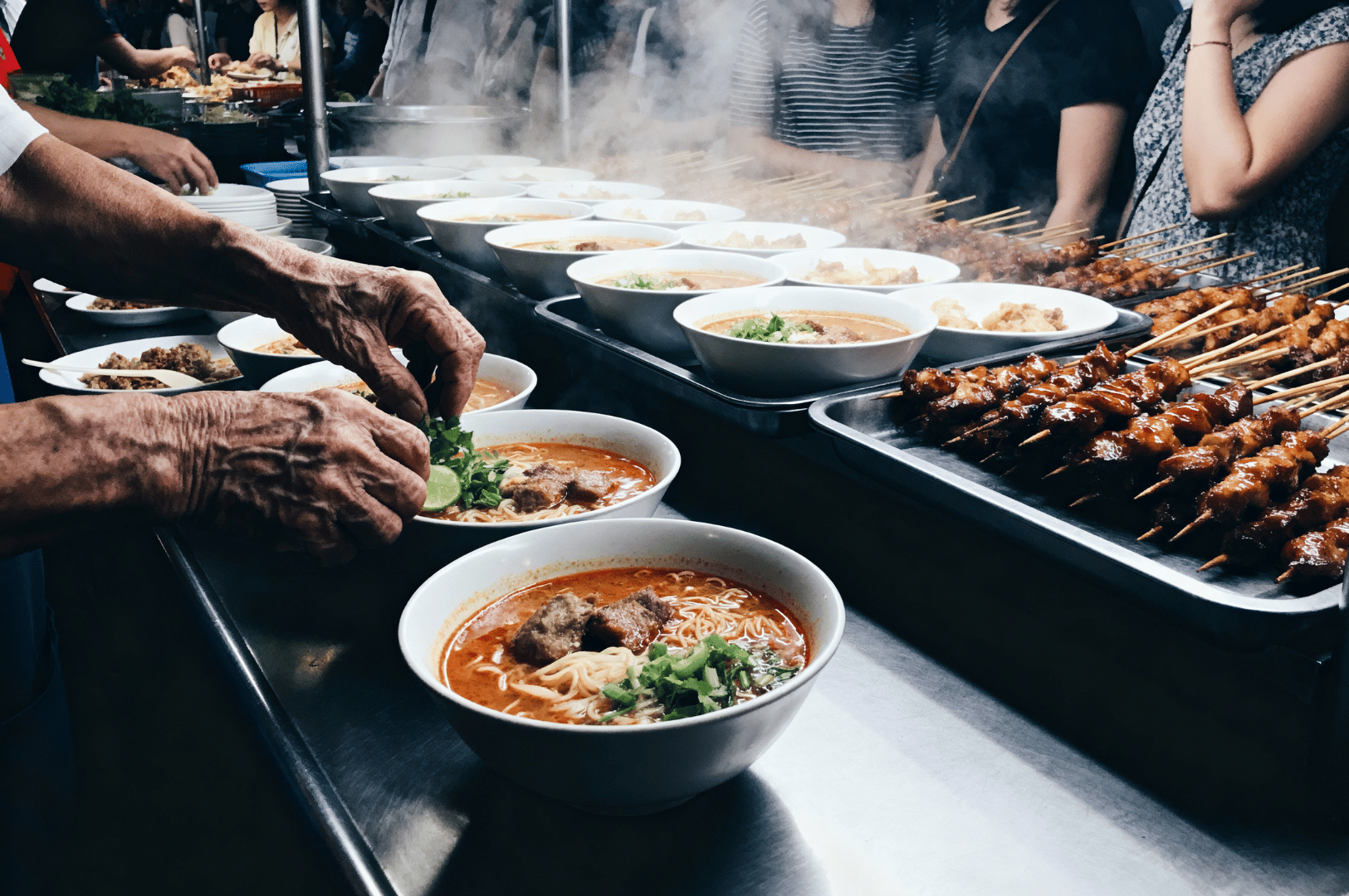 Morning- (4) Hawker stall at breakfast with steaming bowls of noodle soup being garnished, skewers grilling, and a small crowd waiting.
