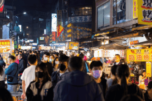 A crowded East Asian night market glows under neon signage and warm stall lights, with masked patrons weaving through narrow lanes lined with sizzling street food—an atmospheric tableau of motion, texture, and ambient contrast ideal for low-light culinary photography.