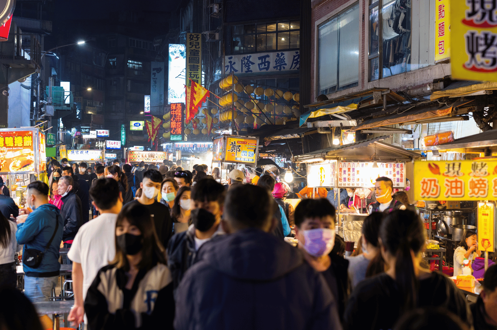 Night-market-Pics- (1) A crowded East Asian night market glows under neon signage and warm stall lights, with masked patrons weaving through narrow lanes lined with sizzling street food—an atmospheric tableau of motion, texture, and ambient contrast ideal for low-light culinary photography.