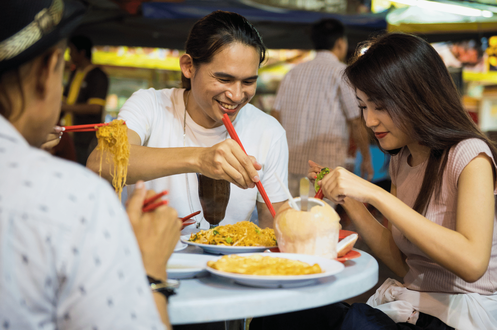 Night-market-Pics- (4) Three friends gather around a street-side table under market lights, sharing noodles and laughter—chopsticks mid-air, coconut drink glistening, and the warm pulse of Southeast Asian street food culture radiating through the scene.