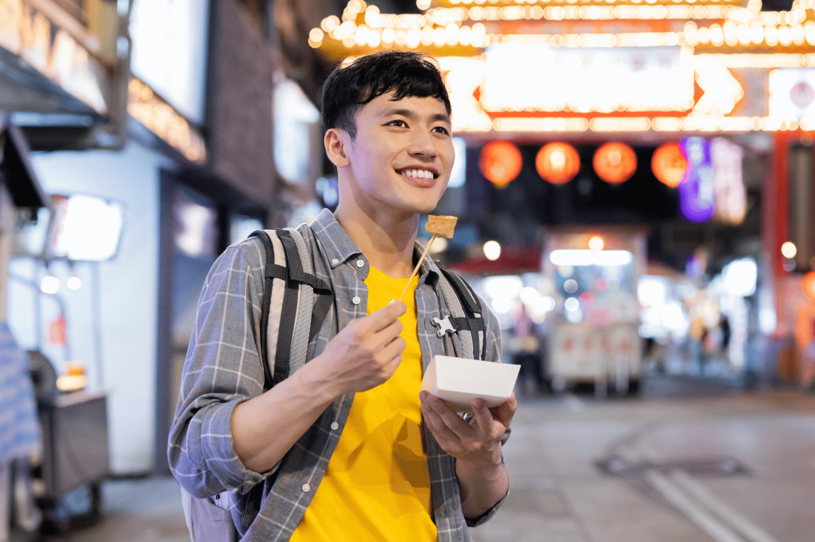 Night-market-Pics- (5) A street food lover stands illuminated by lantern glow, savoring a skewer beside neon-lit signs—his casual layers and backpack hint at a night of urban wandering, while the white container cradles the flavors of a bustling Asian market.