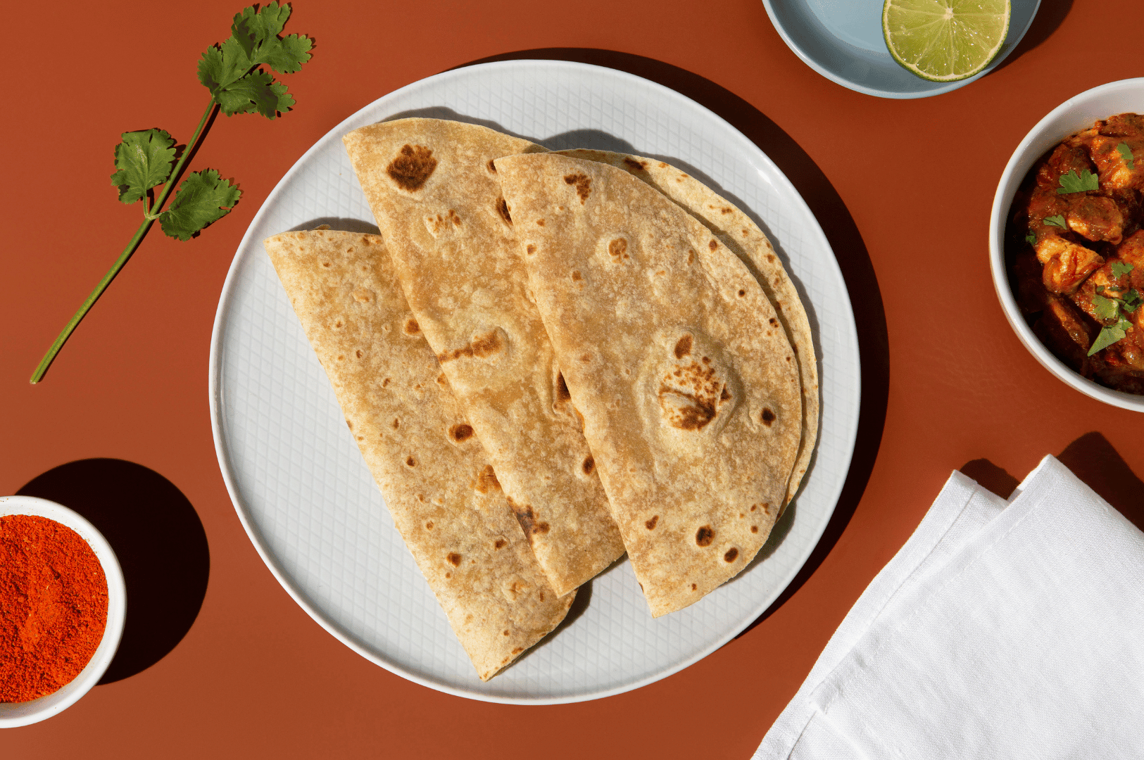Three warm chapatis folded on a white plate, surrounded by chili powder, lime, and a rich curry—an intimate still life of spice and softness, evoking the quiet sensuality behind Singapore’s roti prata obsession.