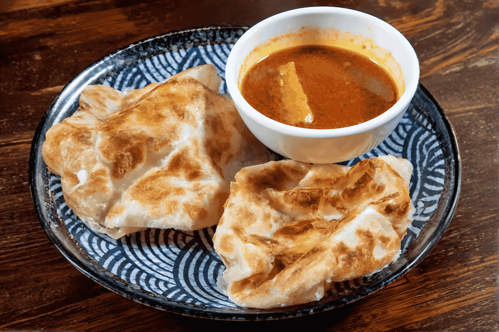 Flaky paratha torn and dipped into a rich paneer curry, the plate’s blue rim framing this tactile ritual—Singapore’s street food scene distilled into one sensual, savory gesture.
