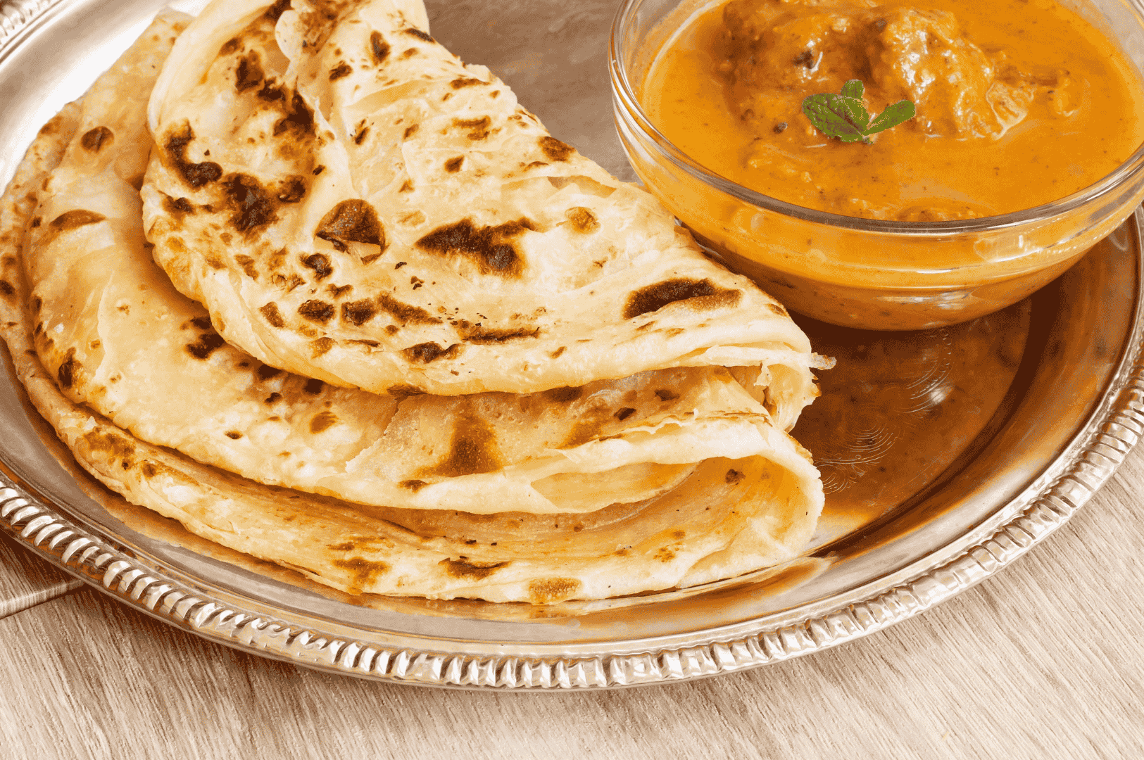 Golden parathas layered on a silver plate beside creamy paneer curry—an elegant, tactile pairing that captures the indulgent stretch and richness of Singapore’s roti prata allure.