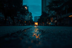 Low-angle shot of a wet, dark city street at night, with a blurred vehicle's red brake lights reflecting in the wet asphalt.