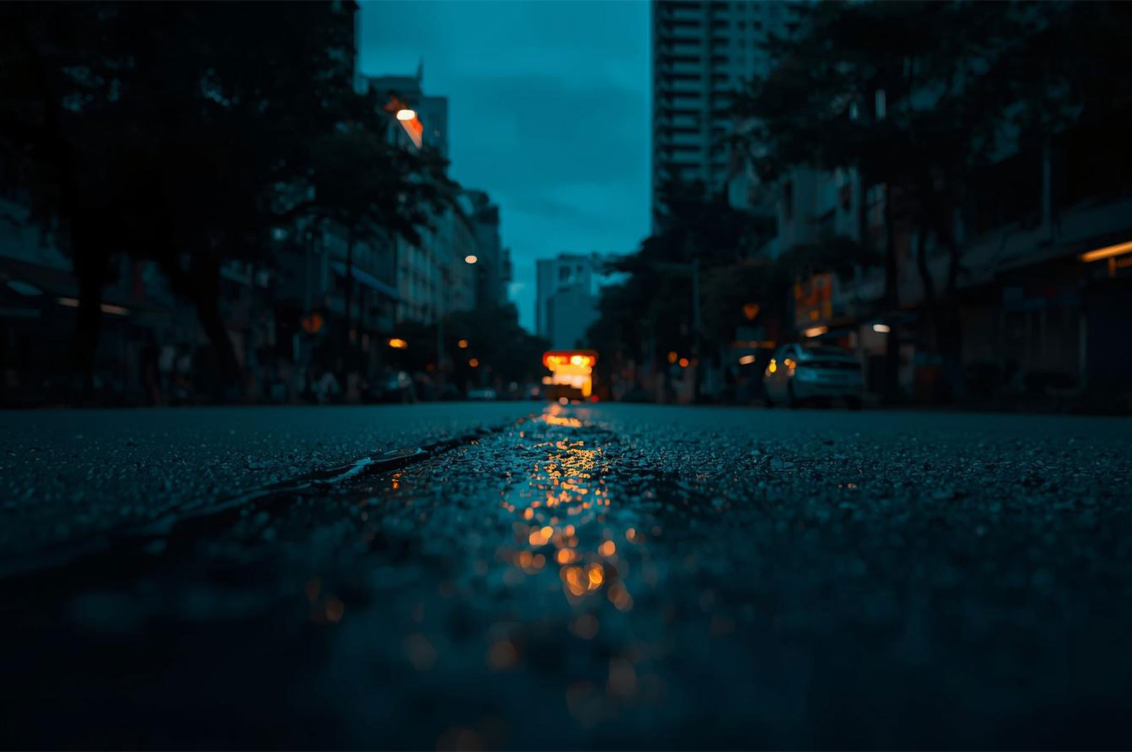 Low-angle shot of a wet, dark city street at night, with a blurred vehicle's red brake lights reflecting in the wet asphalt.