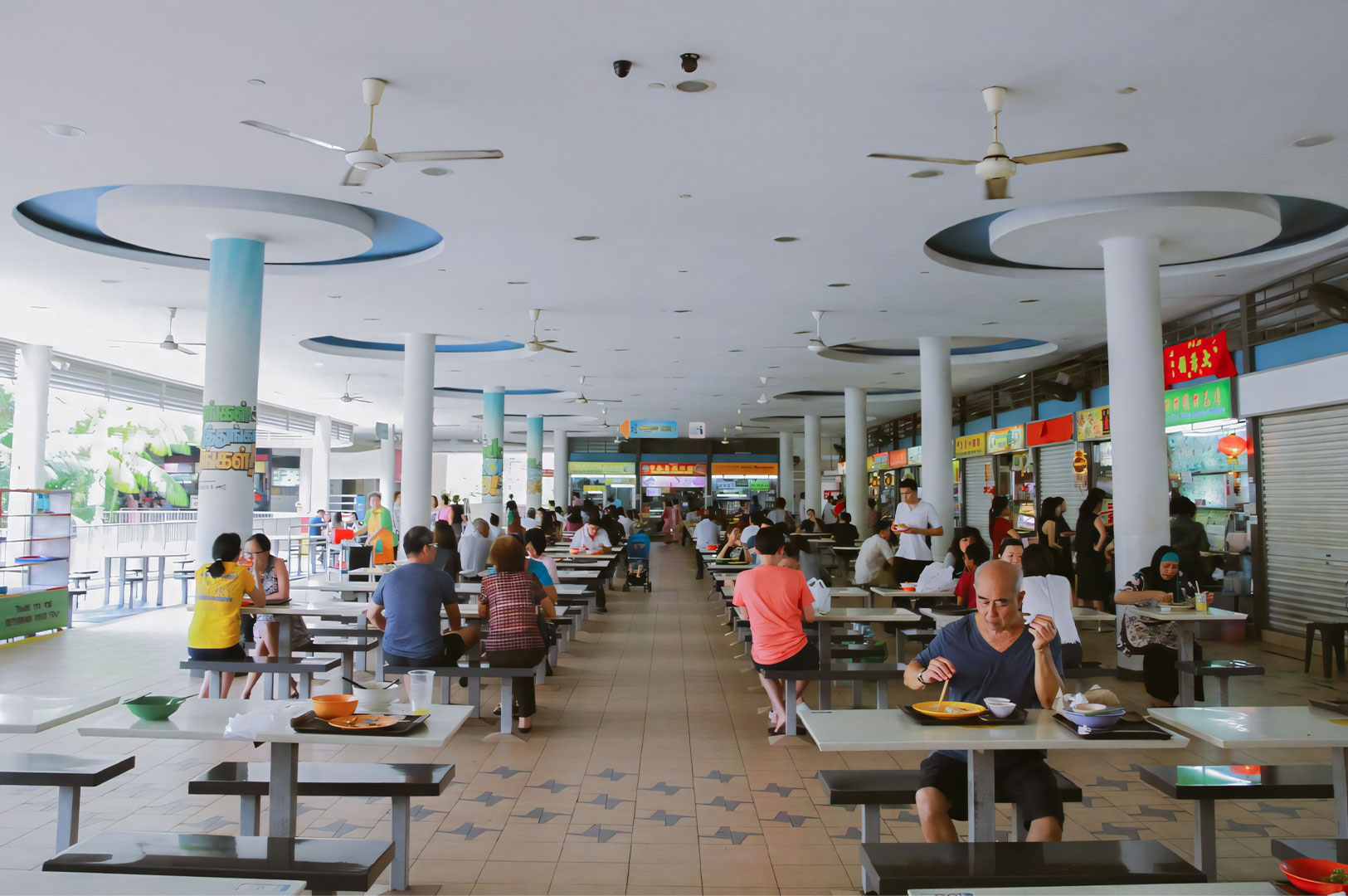 Wide view of a bright, spacious open-air hawker center filled with people sitting at tables eating, showcasing the communal dining environment.