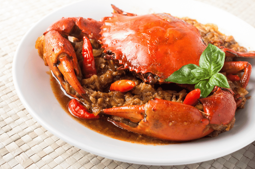Chili-Crab- (1) A whole crab with a brilliant red shell resting on a bed of thick, dark chili sauce, accented by whole red chilies and a fresh green basil garnish on a white plate.