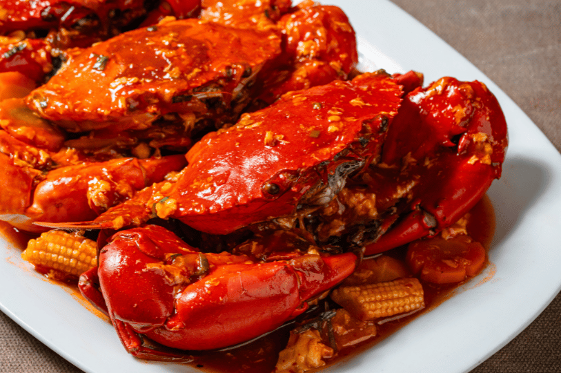 Chili-Crab- (3) A dramatic, side-angle close-up of glossy red crab claws and shells glistening under warm lights, served with a rich sauce containing baby corn and carrots.