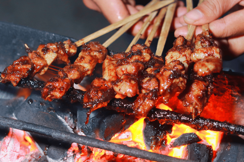 Flame-Satay- (1) A close-up, low-angle shot of several meat skewers grilling over glowing orange charcoal. The meat is deeply caramelized and glistening with marinade, while a person’s hands are visible in the background adjusting the bamboo sticks.