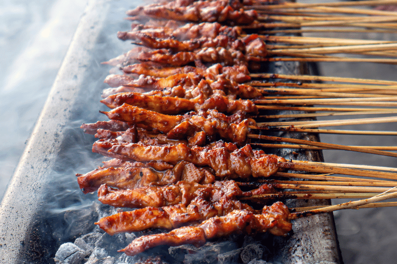 Flame-Satay- (2) A long, perspective view of dozens of satay skewers lined up on a traditional narrow charcoal grill. Thick white smoke rises from the embers, veiling the rows of browning meat in a hazy, atmospheric street-food setting.