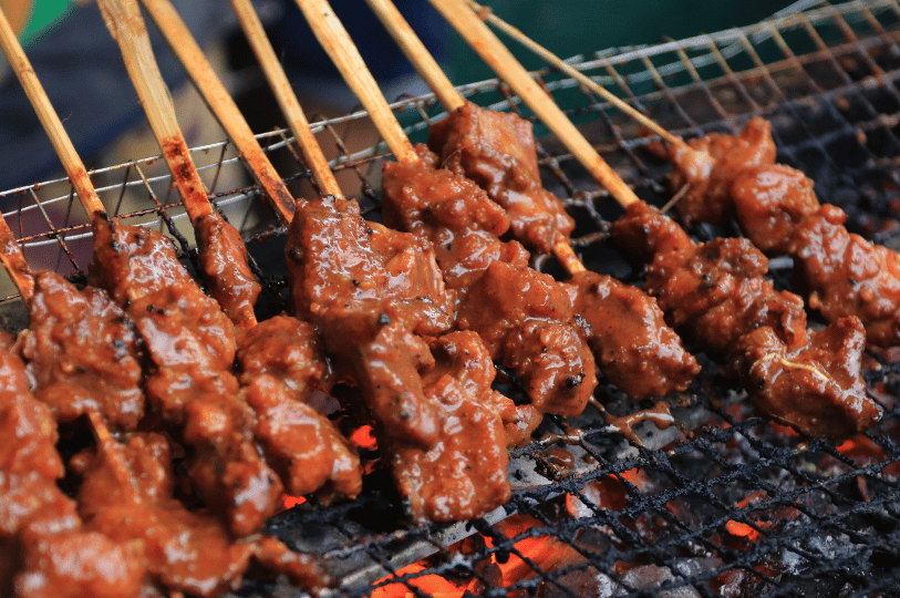 Flame-Satay- (3) Macro photography of satay skewers resting on a wire metal grate. The meat is heavily coated in a thick, textured peanut sauce, with vibrant orange flames and red-hot coals visible directly underneath through the grill mesh.
