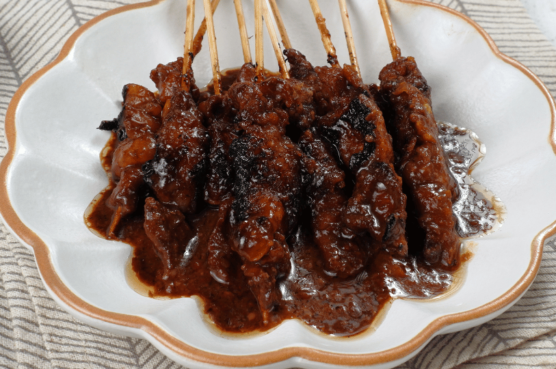 Flame-Satay- (4) A top-down shot of finished satay served on a white floral-shaped ceramic plate. The skewers are piled together, drenched in a rich, dark brown savory sauce that pools at the bottom of the dish.