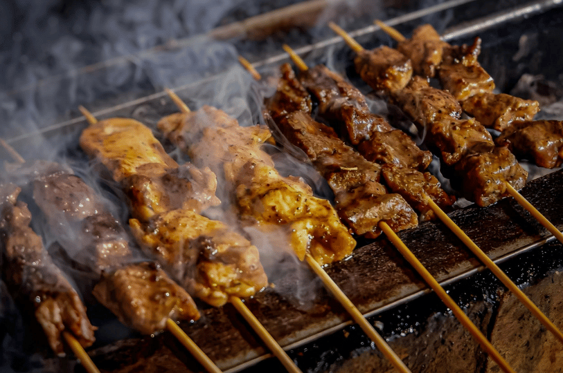 Flame-Satay- (5) A dramatic, moody shot of different varieties of satay—some pale and some dark—grilling side-by-side. Wisps of blue-grey smoke swirl around the skewers, highlighting the contrast between the raw marinade and the charred, cooked edges.