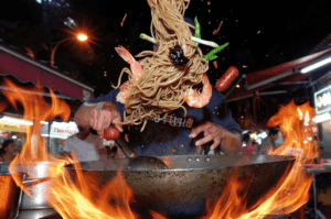 A dynamic action shot of a street food chef tossing noodles, shrimp, and vegetables high into the air from a flaming wok, perfectly freezing the mid-air motion against a vibrant night market background.