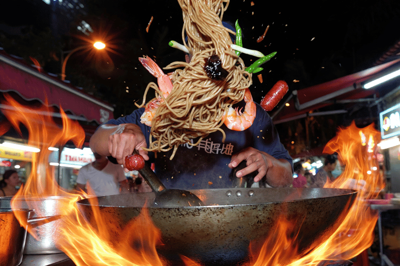 In-Motion- (1) A dynamic action shot of a street food chef tossing noodles, shrimp, and vegetables high into the air from a flaming wok, perfectly freezing the mid-air motion against a vibrant night market background.