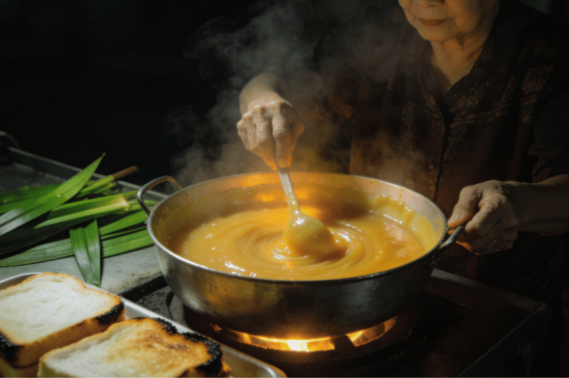 Madam Goh, a kaya toast master, stirs a large, steaming pot of golden-yellow kaya (coconut jam) over a gentle flame, with pandan leaves and slices of slightly burnt toast visible in the foreground.
