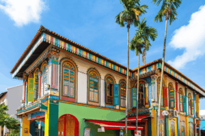A kaleidoscopic heritage shophouse in Little India, Singapore, with ornate stained-glass windows, rainbow-hued shutters, and tropical palms under a bright blue sky—evoking the vibrant spirit of Spice Affair’s culinary journey.