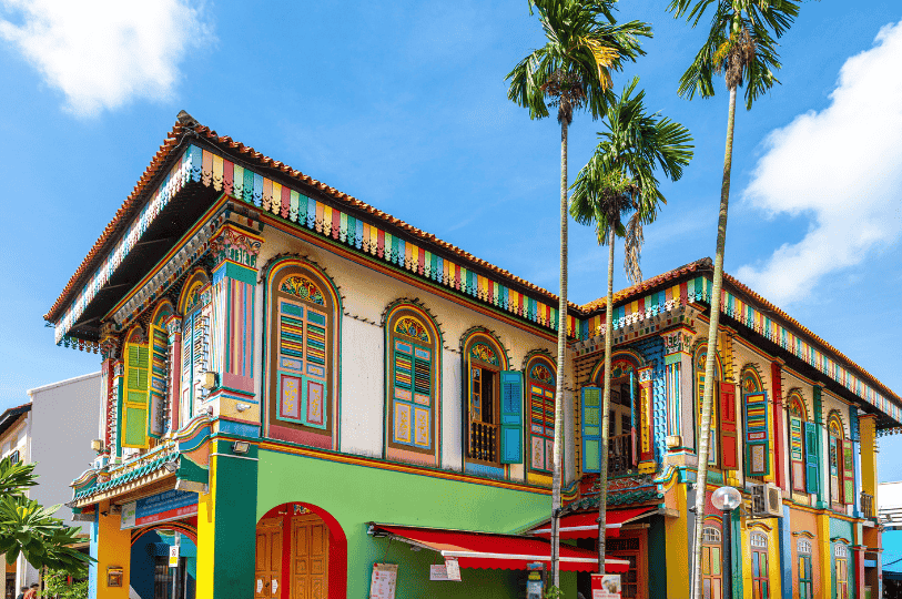 Little-India-1 A kaleidoscopic heritage shophouse in Little India, Singapore, with ornate stained-glass windows, rainbow-hued shutters, and tropical palms under a bright blue sky—evoking the vibrant spirit of Spice Affair’s culinary journey.