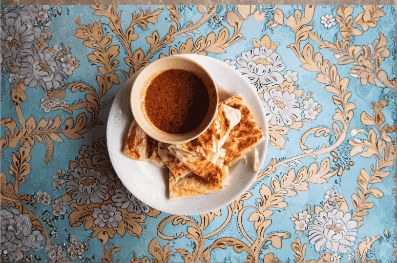 Little-India-2 Golden-brown stuffed flatbreads encircle a bowl of rich curry on a vibrant floral tablecloth—an inviting taste of Little India’s spice-laden street eats.