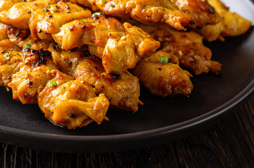 Art-of-the-Tease- (1) A close-up, low-angle shot of glistening chicken satay skewers. The meat is coated in a rich, golden-yellow turmeric marinade with charred edges, topped with black sesame seeds and fresh green garnish against a dark, moody plate.