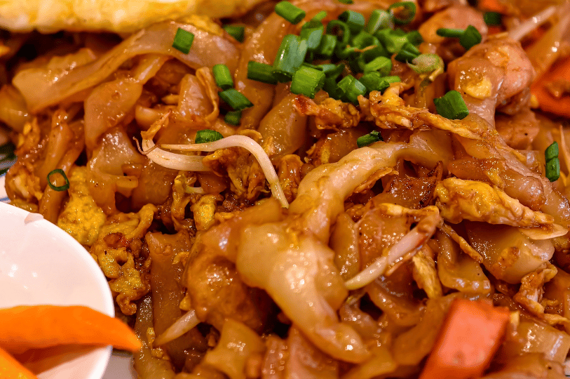 Art-of-the-Tease- (2) A macro view of wide, flat rice noodles (Char Kway Teow) stir-fried to a smoky finish. The image captures the glistening texture of the noodles mixed with bean sprouts, bits of scrambled egg, and sliced green onions.